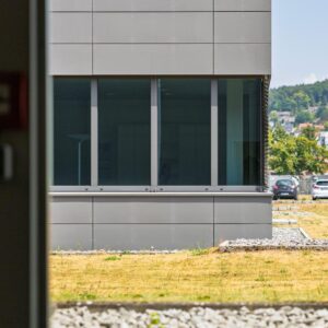 Seitliche Ansicht eines modernen Firmengebäudes mit grauer Fassade, großen Fenstern und Außenanlage.